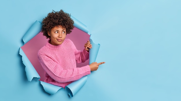 Curly haired young woman gives advice where to go indicates place for advertisement breaks through blue wall wears knitted sweater recommends something