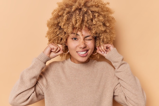 Photo curly haired young woman plugs ears winks eye avoids loud noise doesnt want to hear something unpleasant wears casual jumper isolated over beige background ignores conversation. decibel concept