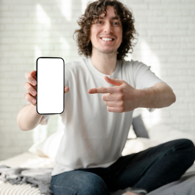 Curly happy guy pointing at the phone and smile sitting on the bed