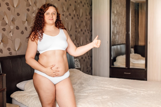 Photo curly happy plus size woman in white underwear stand near bed in bedroom touch thick belly and show good result fat burning treatment of overweight body weight loss program dieting and exercise