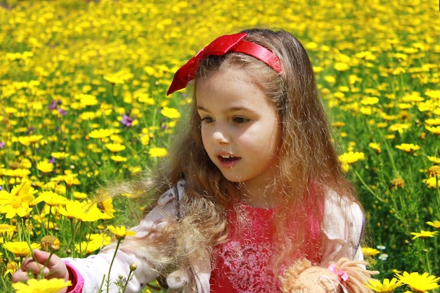 Curly little girl with a red bow in her hair Girl with a dol on a green meadow among yellow flowers