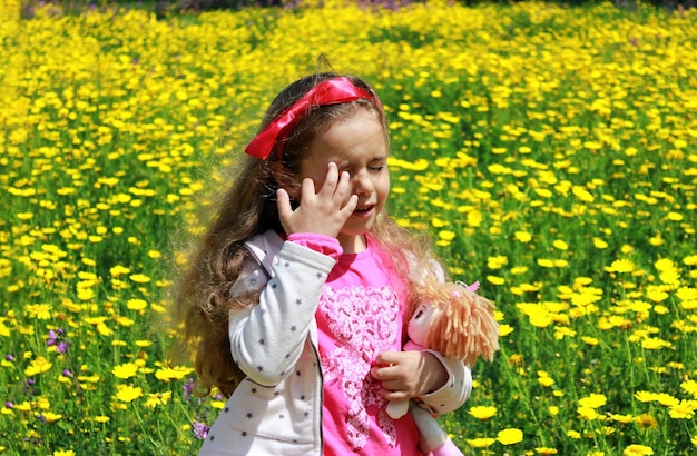 Curly little girl with a red bow in her hair Girl with a dol on a green meadow among yellow flowers