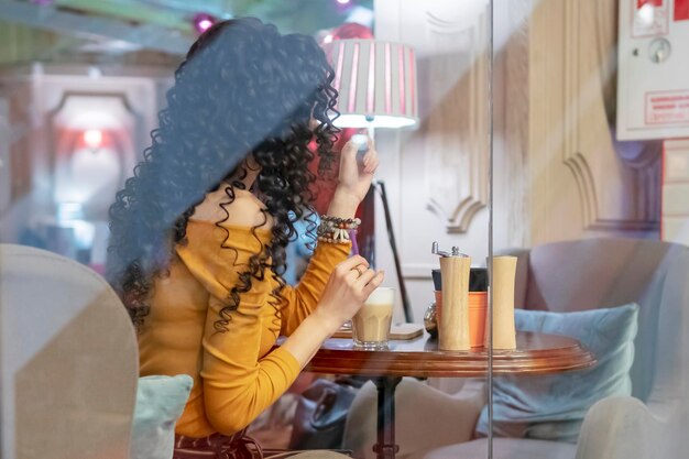 curly lonely woman with a cup of coffee at a table in a cafe behind the glass, rear view