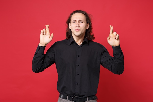 Photo curly long haired man in black shirt posing isolated on red background studio portrait. people lifestyle concept. mock up copy space. waiting for special moment, keeping fingers crossed, making wish.