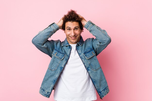 Curly mature man wearing a denim jacket against pink background laughs joyfully keeping hands on head.