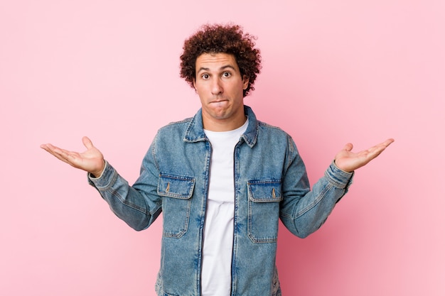 Curly mature man wearing a denim jacket against pink wall confused and doubtful shrugging shoulders to hold a copy space.