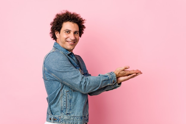 Uomo maturo riccio che porta una giacca di jeans contro la parete rosa che tiene uno spazio della copia su una palma.