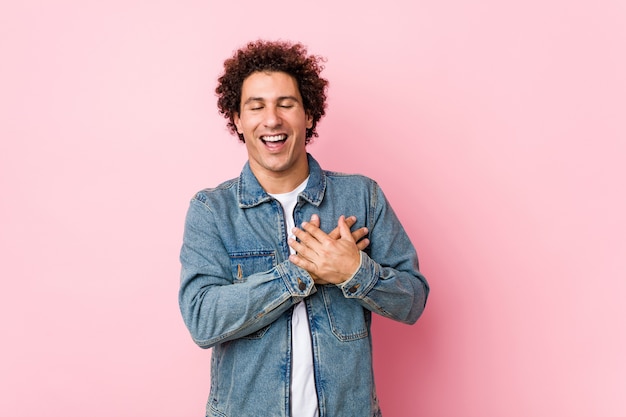 Curly uomo maturo che indossa una giacca di jeans contro il muro rosa che ride tenendo le mani sul cuore, il concetto di felicità.