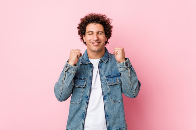 Uomo maturo riccio che indossa una giacca di jeans contro il muro rosa alzando il pugno, sentendosi felice e di successo. concetto di vittoria.