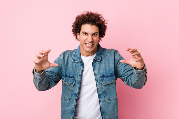 Curly mature man wearing a denim jacket against pink wall screaming with rage.