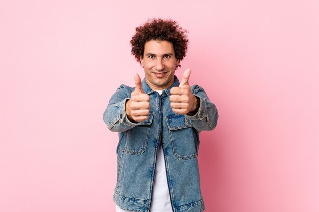 Foto l'uomo maturo riccio che porta una giacca del denim contro il rosa con i pollici aumenta, incoraggia qualcosa, sostiene e rispetta il concetto.