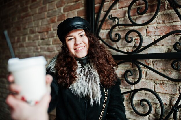 Curly mexican girl in leather cap and plastic cup of coffee at hand walking at streets of city.