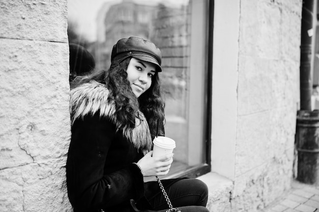 Curly mexican girl in leather cap and plastic cup of coffee at hand walking at streets of city.