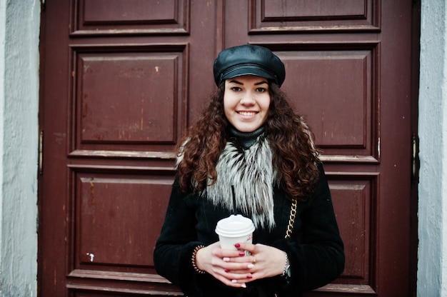 Curly mexican girl in leather cap and plastic cup of coffee at hand walking at streets of city.