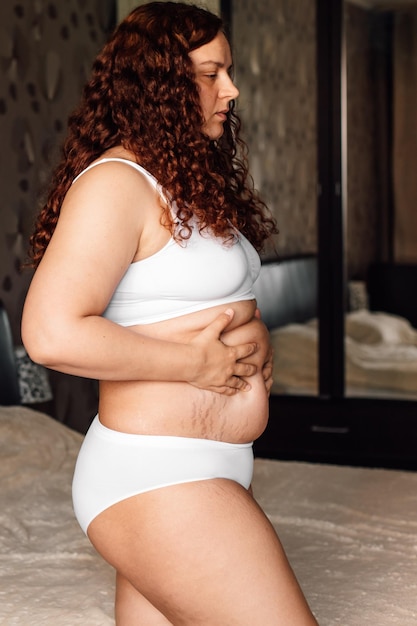 Curly plus size vrouw in wit ondergoed permanent in de buurt van bed in de slaapkamer en drukken op de buik met striae close-up zijaanzicht Vetverbranding behandeling van dikke buik Gewichtsverlies programma dieet