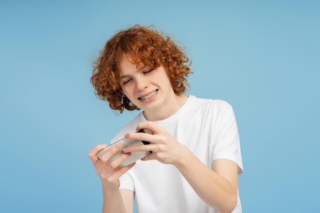 Curly red haired male teenager with braces enjoys gaming on mobile phone