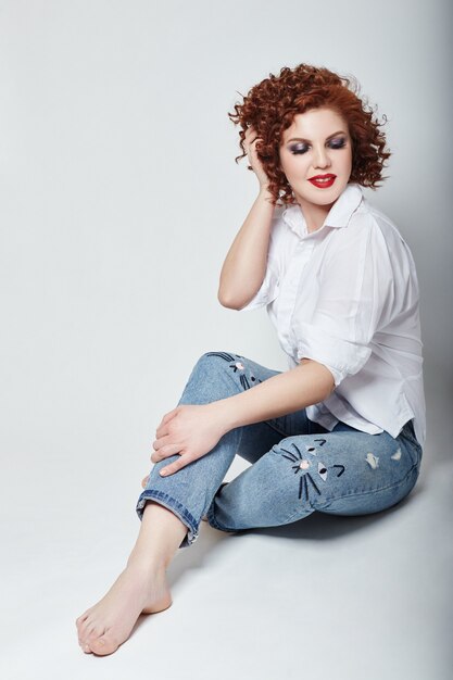 Curly redhead adult sexy woman in white shirt and blue jeans posing sitting in Studio on light background. Russia, Sverdlovsk, 13 March 2018