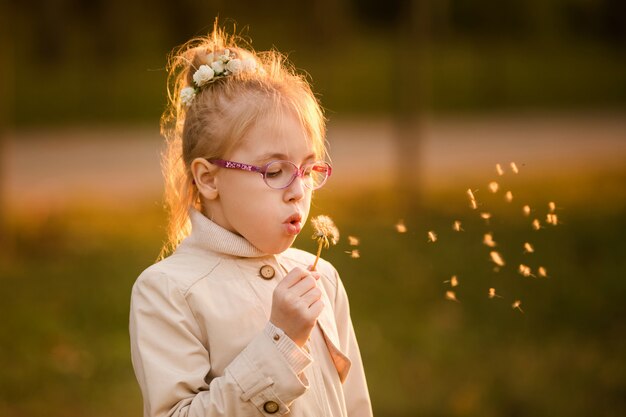 Curly schoolgirl in the glasses and light coat blowing at dandelion in the autumn park on the sunset