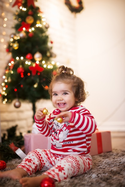 Curly small child woman with ponytail playing with ornaments while sitting on a carpet for Christmas holidays.