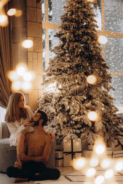 curly smiling woman hugging shirtless muscular boyfriend near christmas tree at home