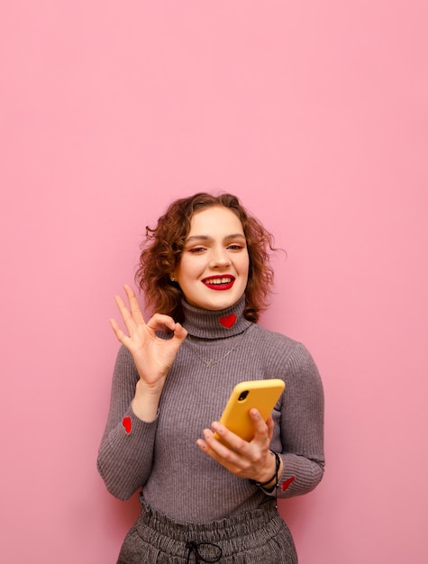 Curly teen girl with smartphone in hand with smile on face looks in camera and shows OK gesture