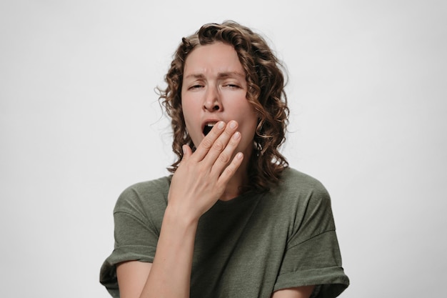 Curly tired sleepy woman yawns and covers mouth with palm, looking bored, stands against white background with blank space for your promotional content