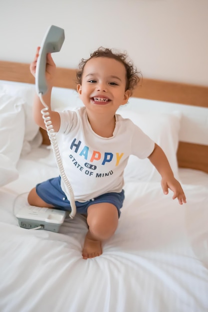 Photo curly toddler boy playing with a wired telephone on the bed