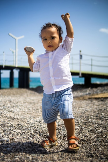 写真 巻き毛の幼児の男の子が青い海の近くの小石のビーチで遊ぶ