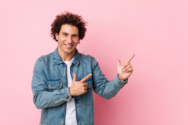 Curly volwassen man draagt een spijkerjasje tegen roze wijzend met wijsvingers naar een kopie ruimte, uiting van opwinding en verlangen.