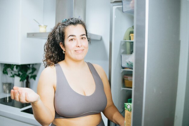 Curly woman doubting about what to eat from the fridge at the kitchen home to lose weight Training clothes Getting fit for the summer conceptBeach body preparation healthy life