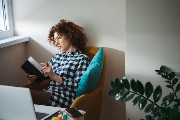 Curly woman in eyeglasses sitting in armchair with laptop on small table aside and taking notes in p...