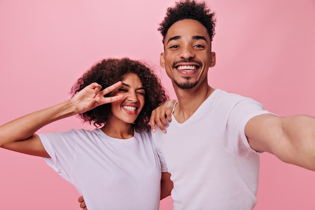 Curly woman showing peace sign and her boyfriend in white T-shirts taking selfie