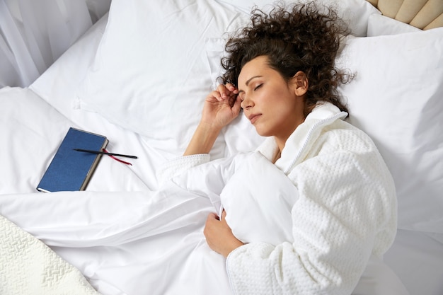 Photo curly woman sleeping in bed with notebook near by wearing bathrobe