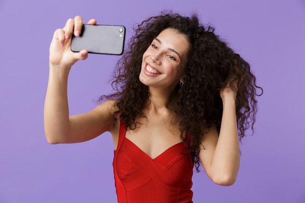 curly woman wearing red dress taking selfie photo on black smartphone, standing isolated over violet wall