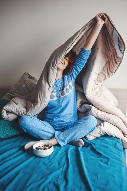 Curly woman who spends her weekend morning in bed under a blanket wearing in blue pajamas healthy me.