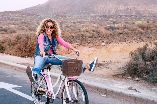 Foto una donna riccia con i capelli biondi e gli occhiali da sole sorride felicemente giocando e divertendosi sulla sua bicicletta sulla strada di campagna