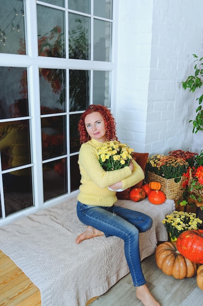 curly woman in yellow sweater sits on window seat in cozy home with autumn decorations, pumpkins