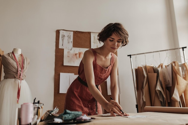 Curly young beautiful brunette woman in linen red dress with belt looks calm works as fashion designer and creates new clothes patterns