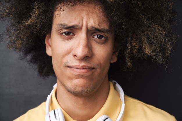 Photo curly young confused sad teenage guy posing over grey chalkboard wall with headphones.