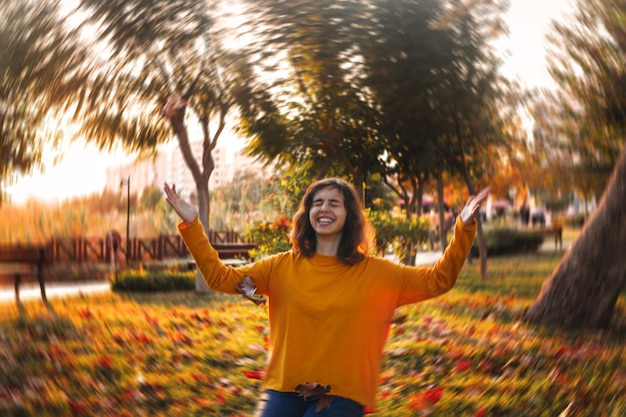 Curly young girl in yellow sweater and jeans sitting on fall grass and throws up dry leaves.