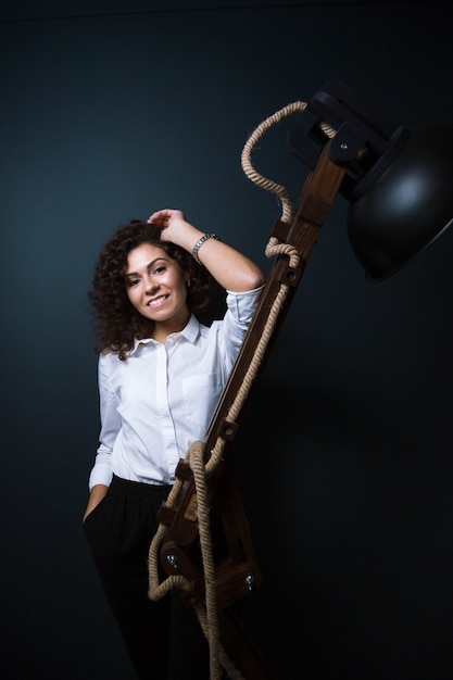 curly young woman in business attire posing by wooden lamp fixture interior element