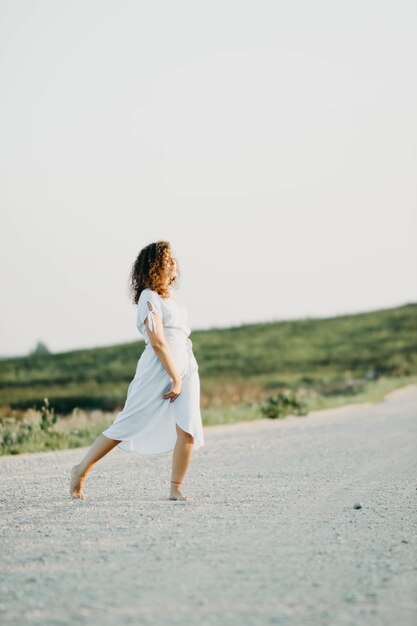 curly young woman dancing romantically in a blue dress on a sandy road at sunset