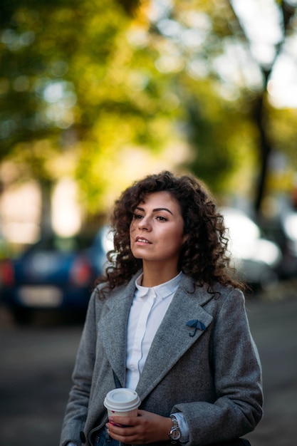 curly young woman in a gray coat walks around the city in autumn