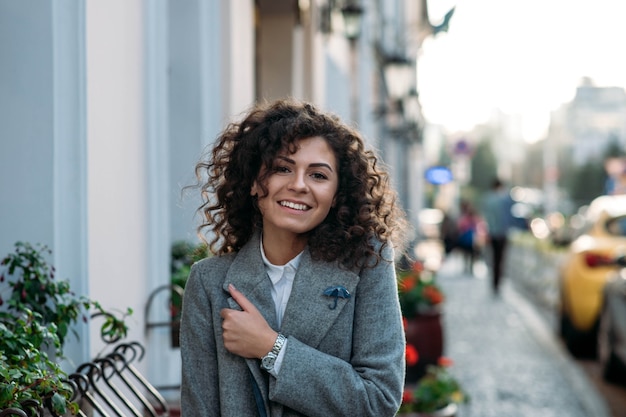 curly young woman in a gray coat walks around the city in autumn