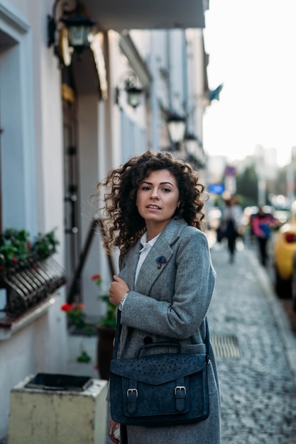 curly young woman in a gray coat walks around the city in autumn