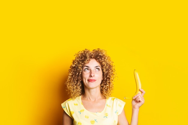 Curly young woman holding a banana pensively looking upwards on a yellow background.