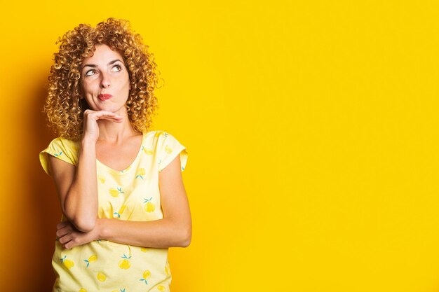 Curly young woman holds her hand under her chin looks thoughtfully upwards on a yellow background