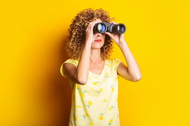 Curly young woman looks through binoculars on a bright yellow surface