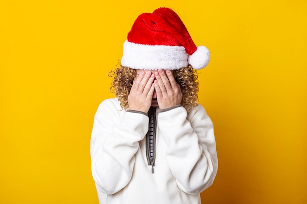 Curly young woman in a santa hat with her palms covering her face on a yellow background.