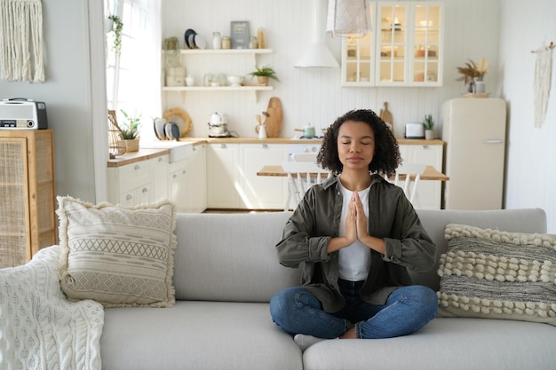 Curly young woman sitting on couch in lotus pose and meditating Wellness and stress relief